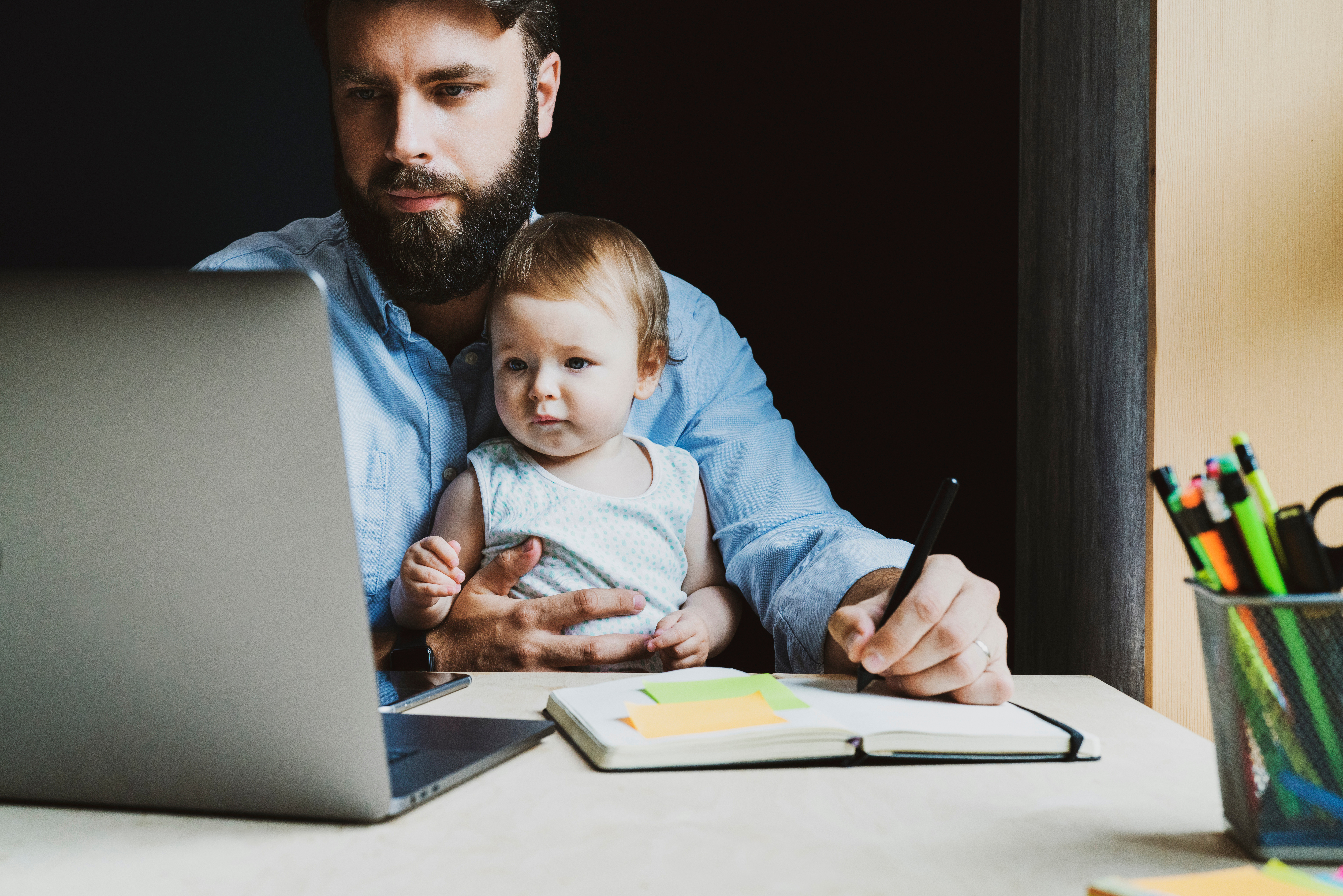 Father with little baby on knees making notes in notebook, looking at laptop. Young man holding child and working on PC. Parent sitting home and getting distant education. Dad doing homebased business